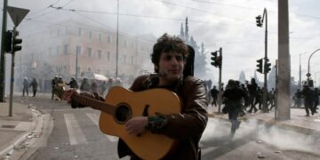 Διαδηλωτής παίζει κιθάρα και τραγουδά την ώρα που δίπλα του πέφτουν μολότοφ και πέτρες